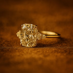 May include: A gold ring with a large, cushion-cut yellow diamond as the centerpiece. The ring has a delicate band and smaller accent stones on either side of the main stone. The ring is set against a warm, brown background.
