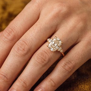 May include: A gold engagement ring featuring a large, rectangular-cut diamond as the centerpiece. The ring has smaller accent diamonds on either side of the main stone. The band is a delicate gold color.