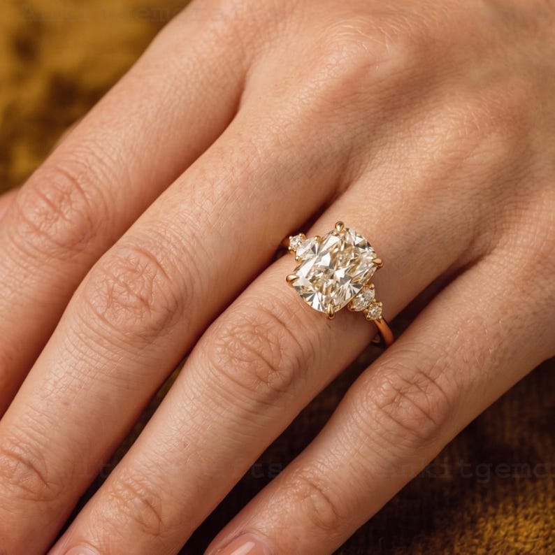 May include: A gold engagement ring featuring a large, rectangular-cut diamond as the centerpiece. The ring has smaller accent diamonds on either side of the main stone. The band is a delicate gold color.