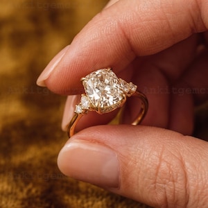 May include: A gold engagement ring featuring a large, cushion-cut diamond as the centerpiece. The ring has smaller accent diamonds on either side and along the band, creating a sparkling effect. The ring is held up against a blurred background.