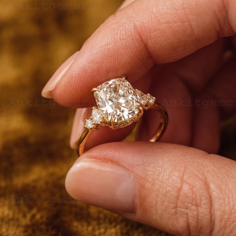 May include: A gold engagement ring featuring a large, cushion-cut diamond as the centerpiece. The ring has smaller accent diamonds on either side and along the band, creating a sparkling effect. The ring is held up against a blurred background.