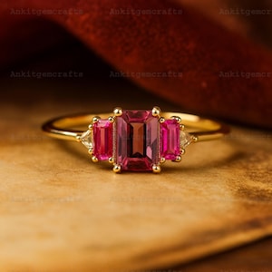 May include: A gold ring featuring a large, rectangular, deep pink gemstone flanked by two smaller, rectangular pink gemstones. The ring also has two triangular, clear gemstones on either side of the pink gemstones.