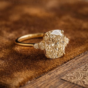 May include: A gold ring featuring a large, rectangular, light-yellow gemstone as the centerpiece. Smaller, round gemstones flank the main stone on each side. The ring is set against a brown, textured background.