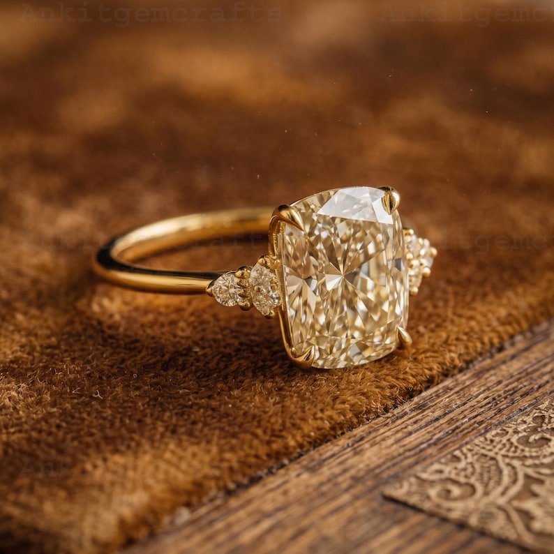 May include: A gold ring featuring a large, rectangular, light-yellow gemstone as the centerpiece. Smaller, round gemstones flank the main stone on each side. The ring is set against a brown, textured background.