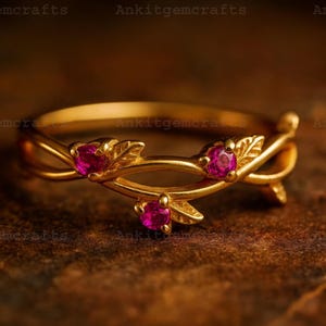 May include: A gold-colored ring with a floral design. The ring features three small, round, pink gemstones and leaf accents. The ring is set against a dark, textured background.