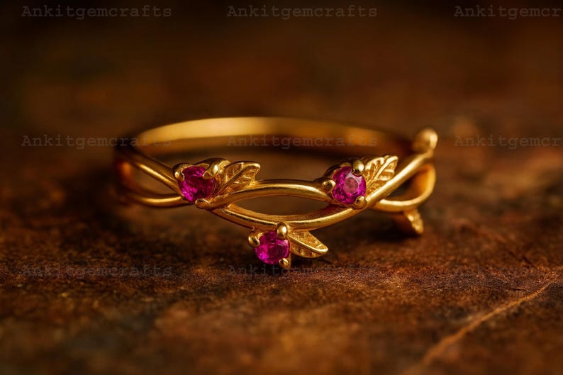 May include: A gold-colored ring with a floral design. The ring features three small, round, pink gemstones and leaf accents. The ring is set against a dark, textured background.