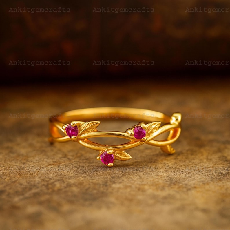 May include: A gold-colored ring with a floral design. The ring features three small, round, red gemstones and leaf-shaped accents. The text "Ankitgemcrafts" is visible in the background.