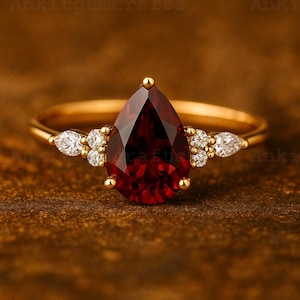以下が含まれることがあります： 中央に大きな洋梨型の深い赤色の宝石をあしらったゴールドリング。リングは、メインストーンの両側に小さなクリアな宝石でアクセントを加えています。バンドはシンプルなポリッシュゴールドです。