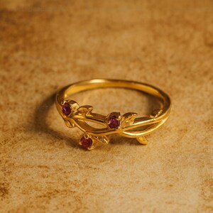 May include: A gold-colored ring with a floral design. The ring features three small, round, red gemstones set within the leaves. The band is a simple, polished gold. The ring is set against a textured, brown background.