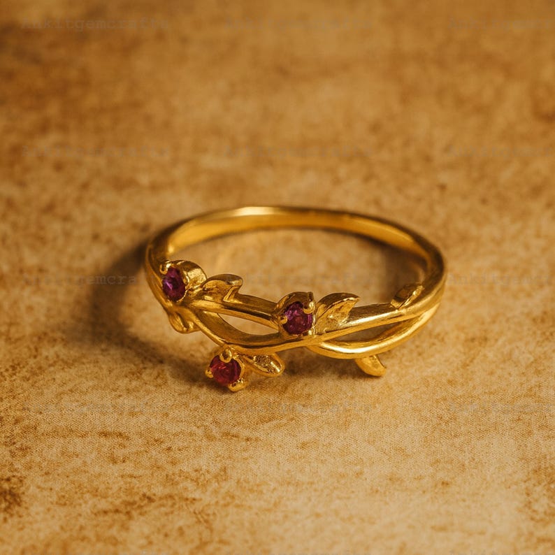 May include: A gold-colored ring with a floral design. The ring features three small, round, red gemstones set within the leaves. The band is a simple, polished gold. The ring is set against a textured, brown background.