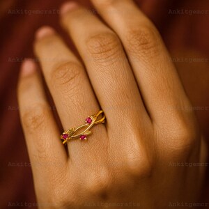 May include: A gold-colored ring with a floral design, featuring three small, round, red gemstones. The ring is on a hand, showcasing its delicate details and craftsmanship. The ring's design includes intertwined bands and leaf-like elements.