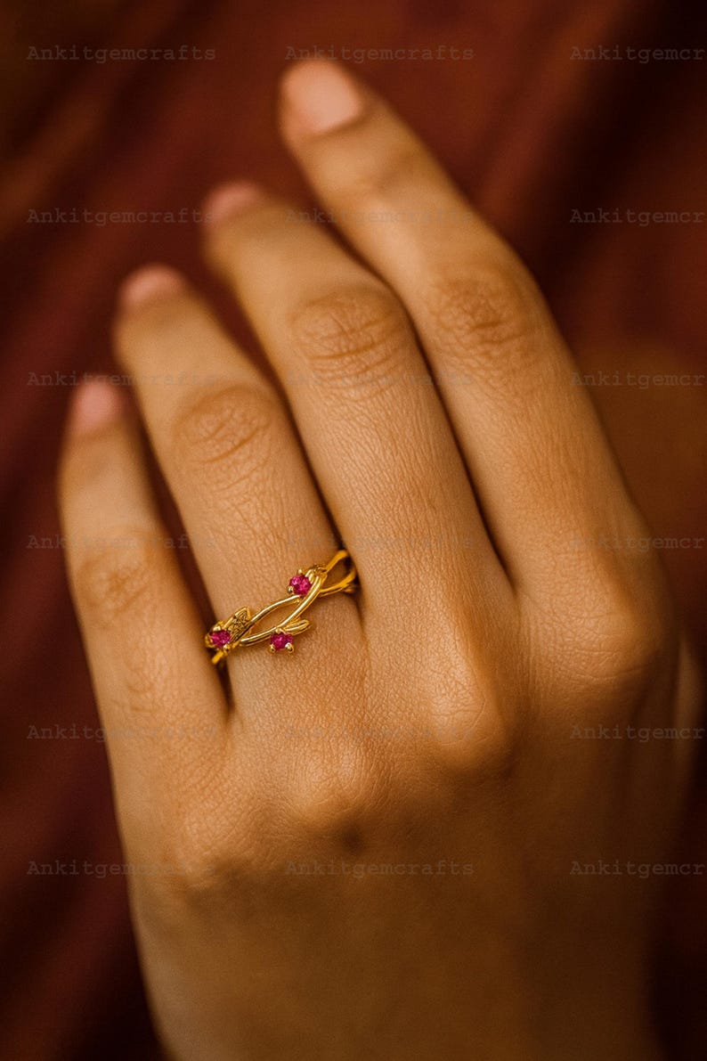 May include: A gold-colored ring with a floral design, featuring three small, round, red gemstones. The ring is on a hand, showcasing its delicate details and craftsmanship. The ring's design includes intertwined bands and leaf-like elements.