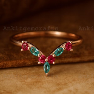 May include: A rose gold ring with a V-shaped design, adorned with vibrant gemstones. The ring features alternating teal and ruby-colored stones, creating a striking contrast. The brand name "Ankitgemcrafts" is visible in the image.