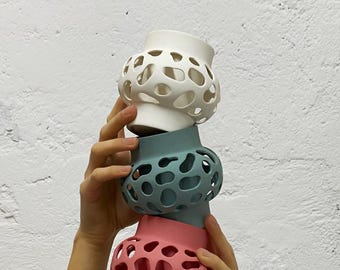 Puede incluir: Tres jarrones decorativos de cerámica en blanco, turquesa y rosa están apilados. Cada jarrón tiene un diseño calado único con agujeros redondeados. Los jarrones son sostenidos por manos sobre un fondo blanco texturizado.