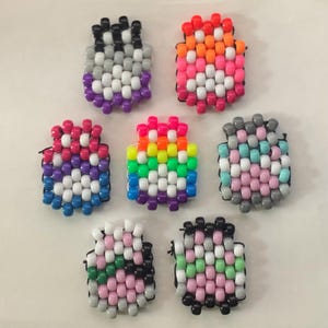 May include: A collection of seven handmade beaded charms in various colors. The charms are made with small, colorful beads strung together to form rectangular shapes. Colors include rainbow, pink, orange, white, black, gray, and purple.