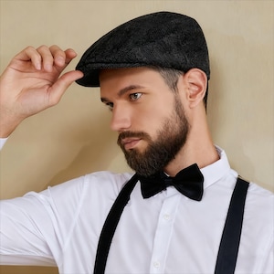 May include: A dark grey herringbone pattern newsboy cap. The cap is worn with a white dress shirt, black bow tie, and black braces. The man in the image has a full beard.