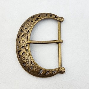 Puede incluir: Hebilla de cinturón de color latón antiguo con un diseño calado ornamentado. La hebilla tiene forma de media luna con una barra rectangular. El intrincado patrón incluye formas florales y geométricas.