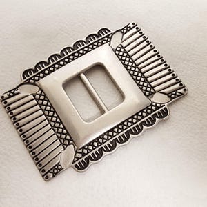 Op de afbeelding: Zilverkleurige rechthoekige gesp met een vierkant midden en een staaf. De gesp heeft een decoratieve geschulpte rand en geometrische patronen. Het ontwerp omvat verticale lijnen en ruitvormige kruisarcering.