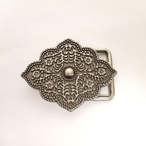 May include: Ornate silver-toned belt buckle with a detailed floral and scroll design. The buckle has a central raised circular element and a rectangular bar for the belt strap. The edges are dotted.