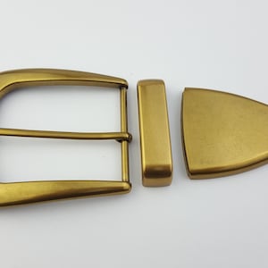 Pode incluir: Um conjunto de três peças de ferragens de cinto de cor latão. O conjunto inclui uma presilha retangular, uma peça final triangular e uma fivela clássica com uma única barra. As peças estão dispostas sobre uma superfície branca.