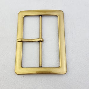 Pode incluir: Fivela de cinto retangular de latão com uma única barra e um pino. A fivela tem cantos arredondados e um acabamento liso e polido. O metal é de uma cor dourada quente.