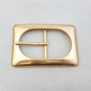 Puede incluir: Hebilla de cinturón rectangular dorada con una abertura ovalada redondeada en el centro. La hebilla tiene un diseño clásico con una sola barra para la correa del cinturón. El metal tiene una superficie ligeramente texturizada.