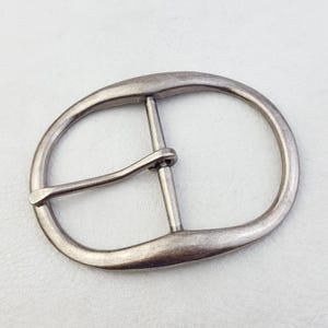 以下が含まれることがあります： 楕円形のアンティークシルバーのベルトバックル。バックルは、中央のバーとベルトを固定するためのプロングを備えたクラシックなデザインです。金属はわずかに風化した外観で、ヴィンテージ感があります。革のベルトに最適です。