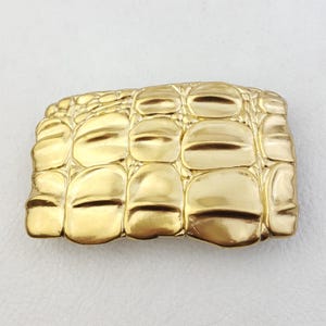 Puede incluir: Hebilla de cinturón dorada con un diseño texturizado de piel de cocodrilo. La hebilla rectangular tiene un borde superior curvo y una serie de formas redondeadas en relieve con hendiduras horizontales, creando un patrón realista de escamas de reptil.