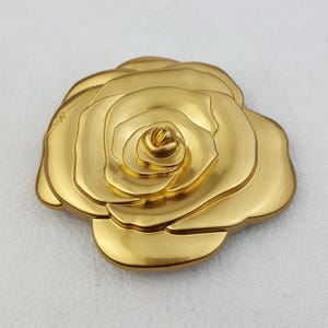 May include: A gold-colored, three-dimensional rose brooch. The brooch is highly detailed, with multiple layers of petals. The surface is smooth and reflective, catching the light. The overall design is elegant and classic, suitable for various occasions.