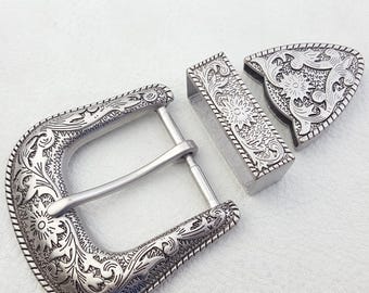May include: A silver-colored belt buckle set with intricate floral and scrollwork designs. The set includes a D-shaped buckle, a rectangular belt loop, and a triangular tip, all with a detailed, embossed pattern. The edges are finished with a rope-like border.