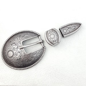 May include: Silver-toned metal belt buckle set with an oval buckle, a rectangular piece, and a pointed end piece. The pieces feature intricate floral and scrollwork designs, with a rope-like border. The set is displayed on a white surface.