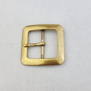 Könnte beinhalten: Eine quadratische, messingfarbene Gürtelschnalle mit rechteckiger Öffnung. Die Schnalle hat einen einzelnen Steg und einen Stift zum Befestigen eines Gürtels. Die Nummer "40" und anderer Text sind auf der Schnalle sichtbar.