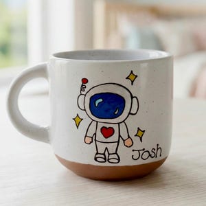 May include: A white ceramic mug with a speckled finish and a brown base. The mug features a hand-drawn illustration of an astronaut with a blue visor and a red heart on the chest, surrounded by yellow stars. The name Josh is written in black ink.
