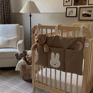 May include: A nursery scene with a light wood crib, a white armchair, and a woven basket filled with stuffed animals. The crib has a brown organizer with a bunny design. The room has framed art and a plaid rug.