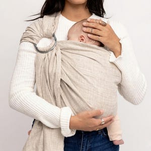 May include: A beige ring sling carrier with a silver metal ring. The carrier is wrapped around a baby, held by a person wearing a white sweater and blue jeans. The baby is wrapped in the sling.