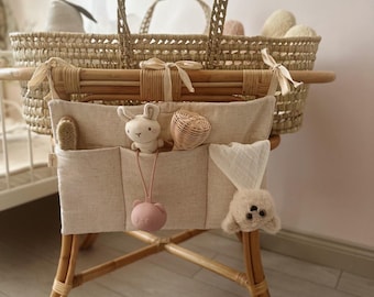 May include: A woven wicker bassinet with a beige fabric organizer, featuring a plush bunny, a small woven basket, and a pink bear-shaped teether. The bassinet is supported by a bamboo frame, creating a cozy and natural aesthetic.