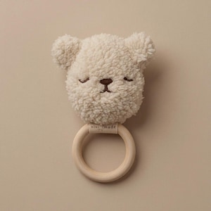 May include: A cream-colored baby rattle shaped like a teddy bear's head with a wooden ring. The bear's face has closed eyes and a small brown nose and mouth. The wooden ring has the text "vivi made" on a small tag.