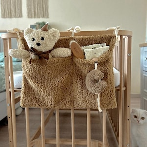 May include: A light brown teddy bear and a matching storage organizer attached to a wooden crib. The organizer has pockets holding a brush, small fabric items, and a plush toy. A white cat sits nearby.