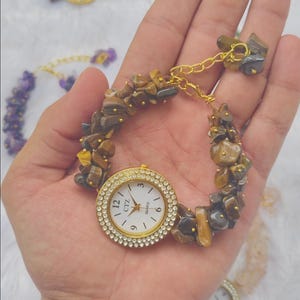 Pode incluir: Um relógio de quartzo dourado com mostrador branco com o texto CTZ e QUARTZ, rodeado por uma luneta de pequenas pedras claras. A bracelete é composta por lascas de pedra irregulares castanhas e cinzentas ligadas por uma corrente dourada.