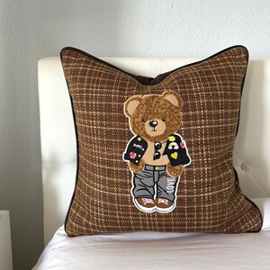 Puede incluir: Cojín decorativo a cuadros marrones con una aplicación de oso de peluche que lleva una chaqueta negra, pantalones grises y zapatos marrones. El oso tiene un parche con la palabra "LOVE" en el zapato. El cojín tiene un borde negro.