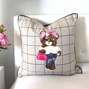 Puede incluir: Un cojín decorativo con un diseño a cuadros en tonos grises y negros. El cojín presenta un oso bordado con gafas de sol, un lazo rosa y un bolso rosa. El cojín tiene un borde negro. Las medidas no son visibles.