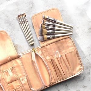May include: A set of makeup brushes with silver handles and soft bristles, arranged with a peach-colored makeup brush case. The case is open, revealing several pockets and a zippered pouch. The brushes are of various sizes and shapes.