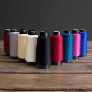 May include: A collection of colorful spools of thread, each with a black base and cap. The thread colors include burgundy, gray, beige, white, black, red, teal, pink, and blue. The spools are arranged on a wooden surface.