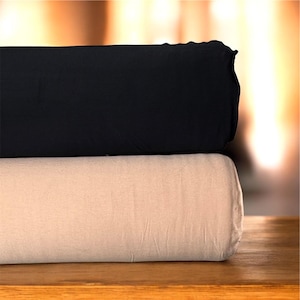 May include: Two folded fabric rolls, one black and one beige, stacked on a wooden surface. The black fabric is on top, with a slightly ruffled edge. The beige fabric is smooth and appears to be a lighter shade.