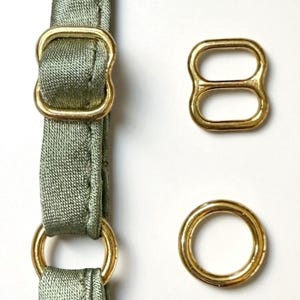 May include: Close-up of gold-colored metal hardware for sewing or crafting. The image shows a gold-colored rectangular buckle, a figure-eight shaped ring, a round ring, and a section of olive green fabric with a matching buckle and ring.