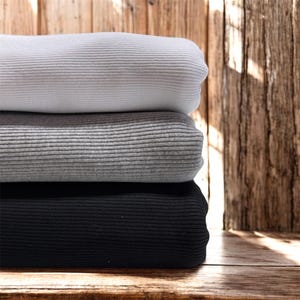 May include: A stack of three folded fabrics in varying shades of gray and black. The top fabric is light gray, the middle is medium gray, and the bottom is black. Each fabric has a ribbed texture, and the stack is placed against a wooden background.