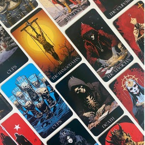 May include: A collection of tarot cards with dark, gothic illustrations. The cards feature images of The Hanged Man, Death, and The Magician, among others. The color palette includes black, red, and blue, with some cards featuring gold accents.