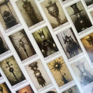 Könnte beinhalten: Ein Kartenspiel mit Tarotkarten und gotischen Illustrationen. Jede Karte zeigt ein detailliertes Bild mit Titeln wie "Stärke", "Tod" und "Der Magier". Die Kunstwerke sind überwiegend in Graustufen gehalten und haben eine Vintage-Ästhetik.