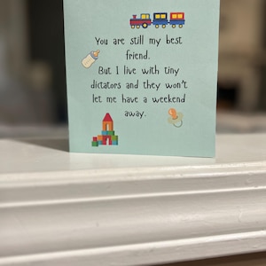 May include: A light blue greeting card with a train, baby bottle, building blocks, and pacifier illustrations. The card reads, "You are still my best friend. But I live with tiny dictators and they won't let me have a weekend away."