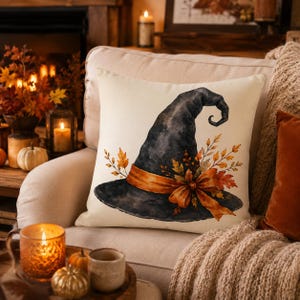 May include: A square throw pillow with an off-white background featuring a watercolor illustration of a black witch hat. The hat is adorned with an orange ribbon tied in a bow and decorated with autumn-colored leaves and small berries.